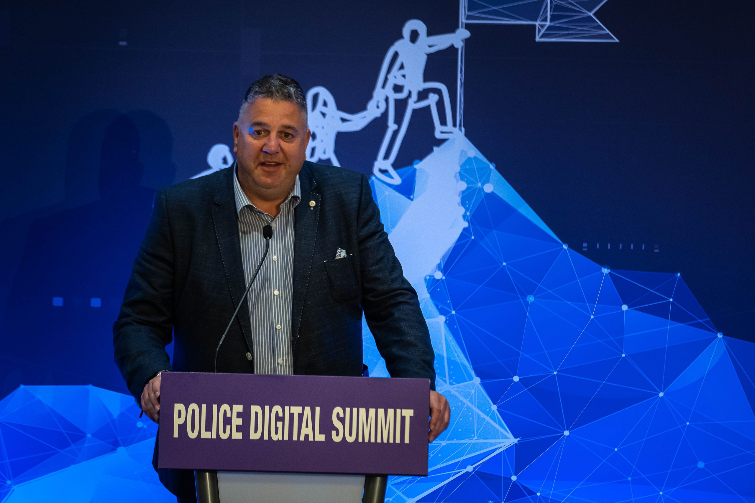 PDS CEO Ian Bell stands in front of a backdrop showing a mountain being scaled by three climbers who are tied together. The lead climber is staking a flag in the summit of the mountain. Ian rests his hand on a lectern which has a purple sign reading "Police Digital Summit" on the front of it. He is dressed in a navy blue blazer over a pale blue shirt.