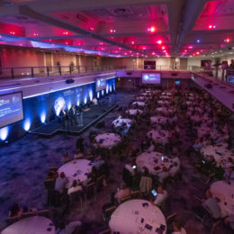 A conference room filled with people seating at their table and looking at the speaker on stage