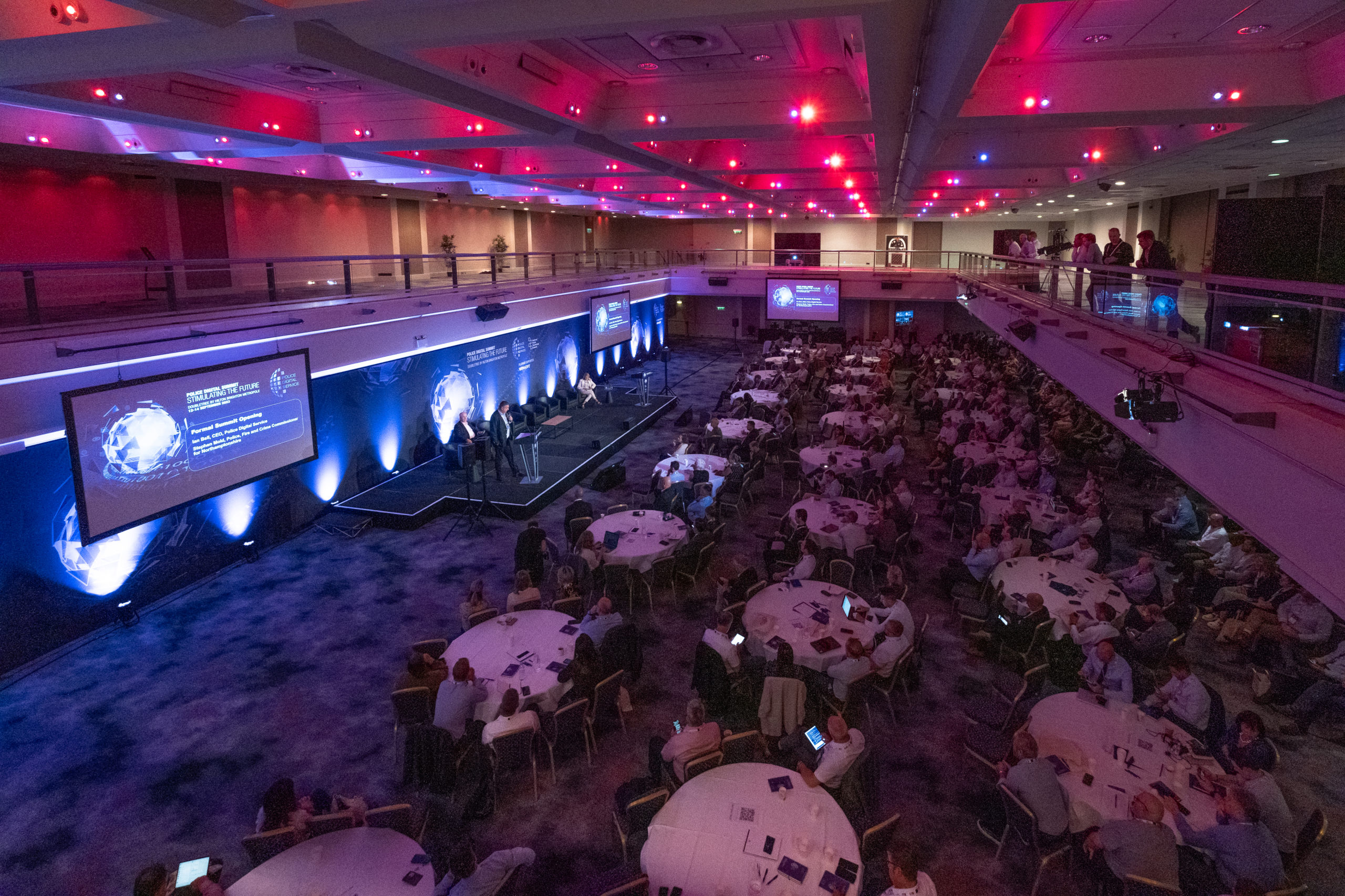 A conference room filled with people seating at their table and looking at the speaker on stage